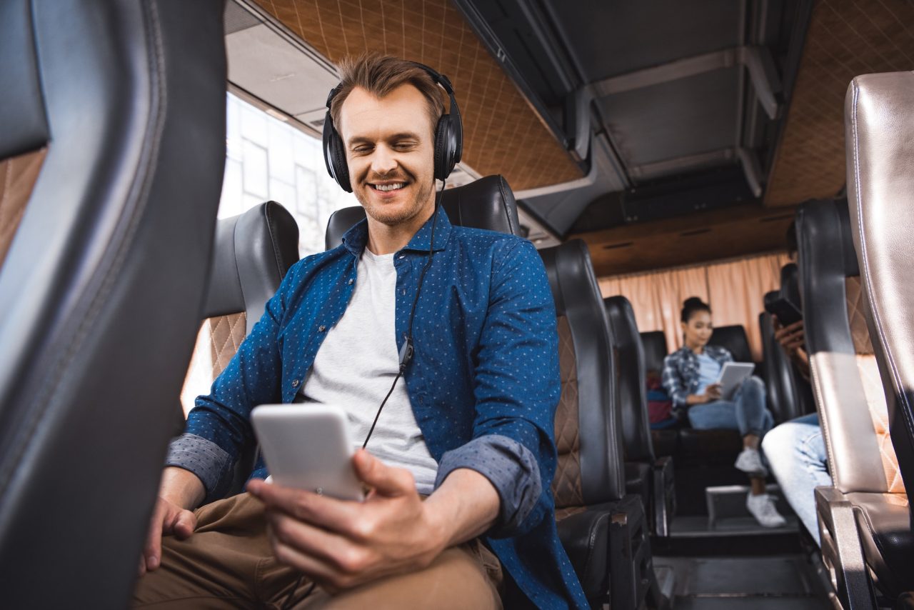 happy adult man in headphones listening music and using smartphone during trip on bus