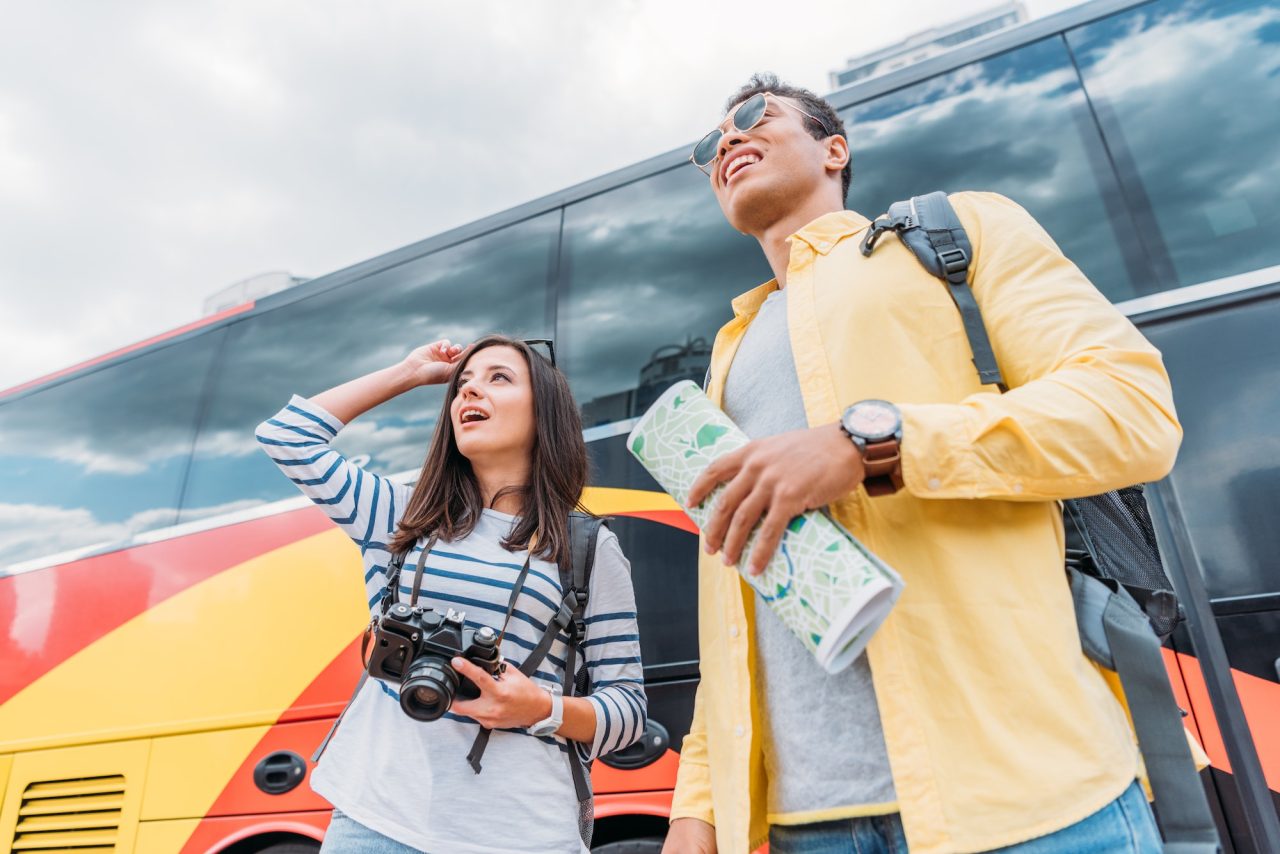 woman holding digital camera and looking away with mixed race man holding map near travel bus